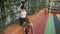 Side view of fit slim sportswoman sitting at mesh fence with green leaves on outdoor court and dancing to music in