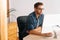 Side view of focused young man wearing stylish glasses working on desktop computer sitting at desk at home office in