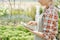 Side view of happy young female worker of greenhouse pointing at display of touchpad at work