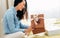 Side view image of happy young woman sit on the bed unpacking the parcel received. Pretty female holding gift box receive order