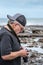 Side view of mature adult photographer with cap looking down at camera on rocks near the sea.