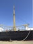 Side view of merchant ship hull and its rigging moored at industrial dock under intense blue sky. Maritime navigation and