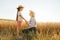 side view of mother and daughter in hat holding hands in golden wheat field.