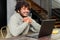 Side view at optimistic Indian man wearing hoodie sitting at desk and using laptop