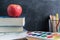 Side view over a wooden desk with school supplies on it. Bucket with colored pencils, notebooks, books, apple, paints. Back to sch