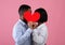 Side view of passionate black lovers kissing behind red paper heart, expressing their love on pink studio background