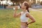 Side view photo of dark haired woman wearing white top drinking water from the bottle, keeps hand on hip, feeling thirsty during