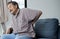 Side view photo of suffering senior man feeling lower back pain, sitting on comfortable sofa in incorrect pose