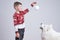 Side view portrait of the child boy in red santa sweater that shows white new year`s toy to his samoyed dog, grey wall