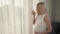 A side view of a pregnant blonde standing by the window covered by the tulle. She caresses her belly, thinks of