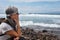 Side view of a senior woman sitting and thinking on the beach. Horizon over water in background. Cloudy sky and waves for surf