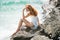 Side view of serene woman sitting on sandy rocky beach against blue waves. Attractive young woman wearing sexy summer