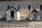 Side view of a small church with wooden door, stone walls, windows, tombs, a cross with Jesus surrounded by a metal fence