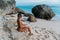 Side view tanned european girl in bathing suit sitting on rocky beach of ocean