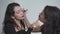 Side View Of Two Pretty Colombian Girls Applying Makeup With Each Other While Sitting On Bed Isolated Against White Wall