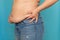 Side view of unrecognizable fat plump woman standing in blue jeans, squeezing excess naked waist on blue background.