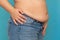 Side view of unrecognizable plump woman in blue jeans putting hands on waist, showing excess belly on blue background.
