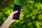 Side view of a woman hand holding a modern slick smartphone while dialing with her thumb against summer green background