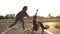 Side view of a woman with her hands raised up sitting on a longboard while her friend is pushing her behind and running