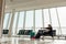 Side view of woman passenger with dark hair wearing black clothes, sitting on chair seat in transit lounge of airport.