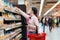 Side view of young Caucasian woman reaches hand to top shelf, holds cellphone and cart. Shelves with food in background