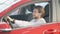 Side view of young confident man sitting in red car on driver\'s seat turning to camera making victory gesture and