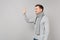 Side view of young man in gray sweater, scarf waving and greeting with hand as notices someone on grey wall