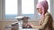 Side view of a young muslim woman in a pink hijab typing on a laptop among the stacks of books