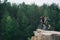 side view of young trial bikers standing on rocky cliff with blurred pine forest on background and looking