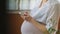 Side-view of youthful woman pregnant women in white t-shirt tapping on smartphone staying in front of the wooden window