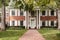 Sidewalk leads to upscale house with white shutters and columns and rocking chairs on porch through tall trees and green grass sca