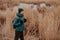 Sideways shot of active male dressed in warm jacket and hat, carries rucksack, has walk in rural area, poses near lake background