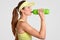 Sideways shot of exhausted female tennis player drinks cold water from bottle, takes break after long match, dressed in tennis cap
