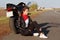 Sideways shot of pensive young experienced female biker wears leather jacket, white sneakers, sits near motorbike on asphalt, enjo