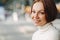 Sideways shot of pleasant looking woman with make up, tender smile, dressed in white turtleneck sweater, stands against blurred