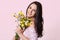 Sideways shot of pleased cute lady with gentle toothy smile, dressed in fashioable dress, holds flowers closely to herself, enjoys