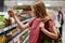 Sideways shot of pretty young femake customer holds canned goods in glass container, stands in food store, dressed in casual cloth