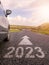 Sign 2023 on small asphalt country road and a side of a car and arrow. Travel and explore concept. Warm sunny day, sun flare.