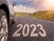 Sign 2023 on small asphalt country road and a side of a car. Travel and explore concept. Warm sunny day, sun flare. Green hill on