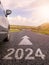 Sign 2024 and directional arrow on a small asphalt road and a side of a car, cloudy sky and country side in the background.