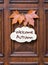 Sign with lettering words Welcome Autumn decorated with two orange maple leaves hanging on dark wooden entrance door.