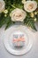 Siingle white plate with a gift box on top and white roses above in a table setting for a ceremony