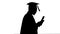 Silhouette African american male student in graduation robe texting on the phone while walking with his diploma.