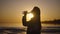 Silhouette of a beautiful woman on the background of the setting sun. A young woman drinks water from a bottle.