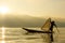 Silhouette Burmese fisherman wearing a hat standing at the back of a boat rowing with legs in the morning