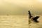 Silhouette Burmese fisherman wearing a hat standing at the back of a boat rowing with legs in the morning