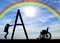 Silhouette of a child disabled girl on a swing next to a wheelchair on a background of the sky with a rainbow