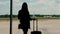 Silhouette of elegant business woman with hand luggage, looking through the window to runway and departing plane, at the