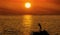 Silhouette of a fisherman on the background of round brilliant setting sun standing on a rock on the sea coast among