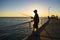 Silhouette of fisherman with hat and fish rod standing on sea dock fishing at sunset with beautiful orange sky in vacations relax